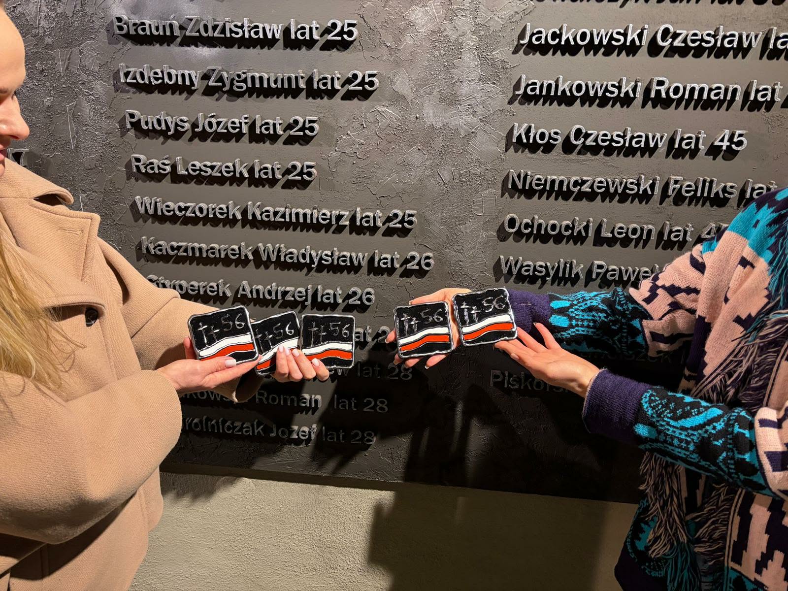 Dwadzieścia kwadratowych, metalowych plakietek. Na czarnym tle srebrne dwa krzyże, cyfra 56 oraz flaga Polski. Flaga Polski jest biało-czerwona.
