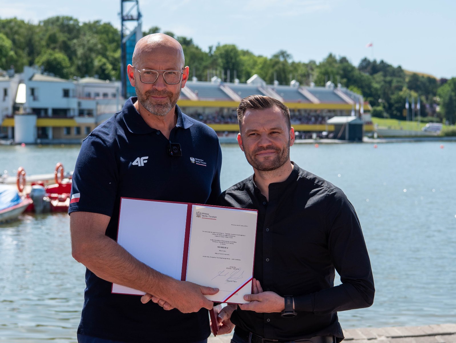 Dwaj mężczyźni stoją obok siebie nad brzegiem jeziora i wspólnie trzymają otwartą, czerwoną teczkę z oficjalnym dokumentem. Mężczyzna po lewej stronie jest wysoki, łysy, nosi glasses i ma na sobie ciemnogranatową koszulkę polo z białym logo "4F" oraz polską flagą na rękawie. Mężczyzna po prawej ma krótkie, ciemne włosy, lekki zarost i jest ubrany w czarną koszulę.