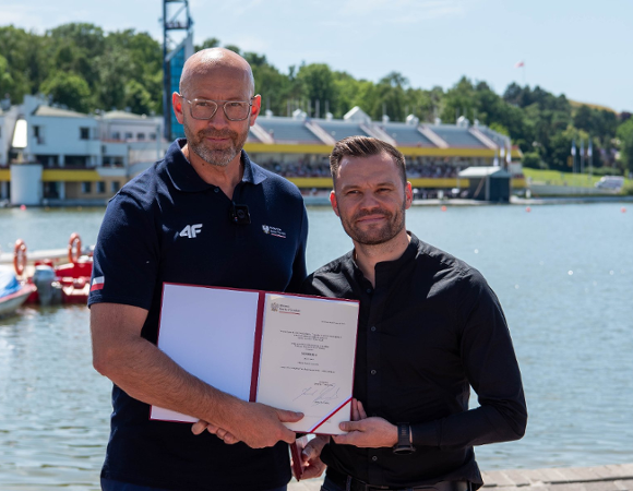 Dwaj mężczyźni stoją obok siebie nad brzegiem jeziora i wspólnie trzymają otwartą, czerwoną teczkę z oficjalnym dokumentem. Mężczyzna po lewej stronie jest wysoki, łysy, nosi glasses i ma na sobie ciemnogranatową koszulkę polo z białym logo "4F" oraz polską flagą na rękawie. Mężczyzna po prawej ma krótkie, ciemne włosy, lekki zarost i jest ubrany w czarną koszulę.