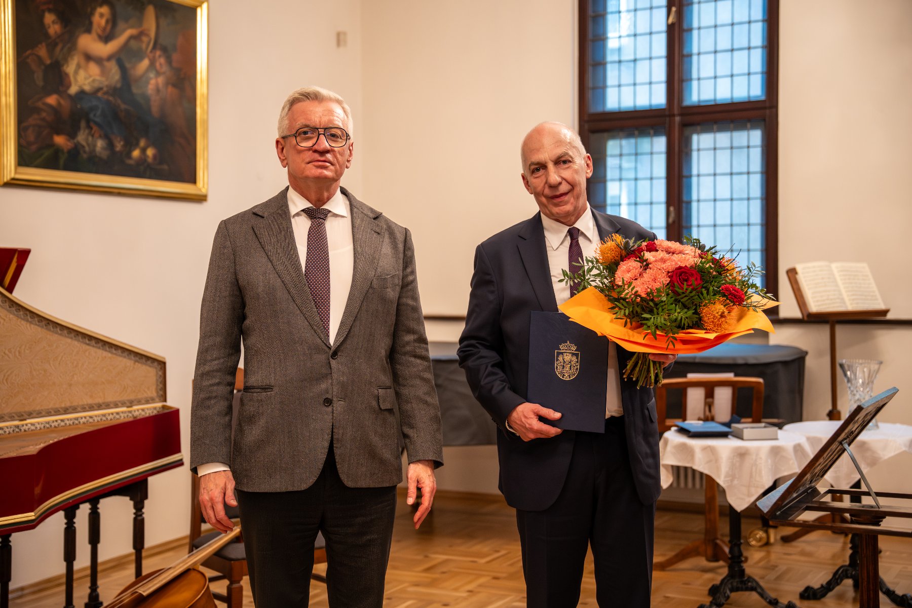 Dwaj mężczyźni w garniturach stoją obok siebie w sali z instrumentami dawnymi. Mężczyzna po prawej trzyma duży bukiet kolorowych kwiatów oraz ciemnoniebieską teczkę z godłem.