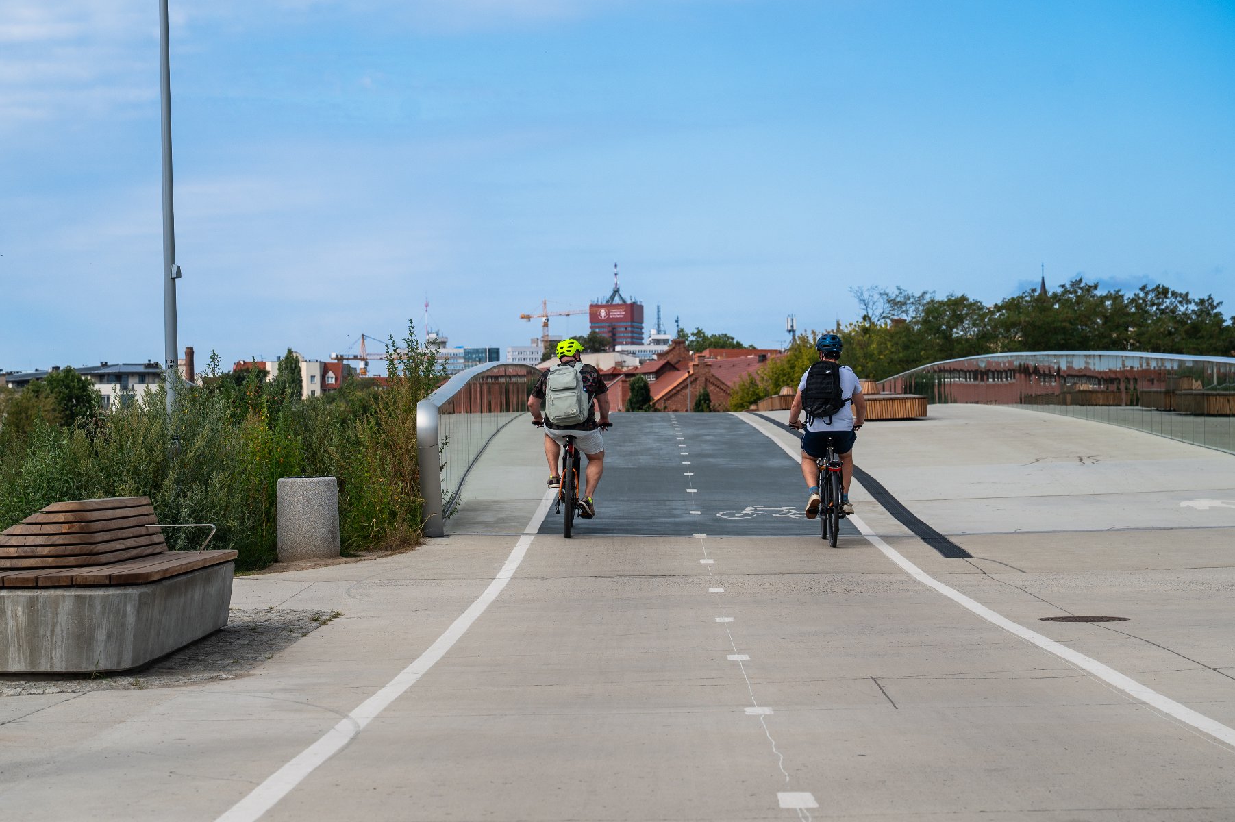 Dwaj rowerzyści z plecakami jadą obok siebie wyznaczonymi pasami na nowoczesnej, betonowej kładce. Po lewej stronie widoczna jest nowoczesna ławka oraz zieleń, a w oddali zarysy budynków miasta i dźwigi budowlane pod jasnym niebem.