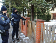 Dwie funkcjonariuszki Straży Miejskiej w granatowych mundurach i czarnych czapkach, stojące na zaśnieżonym chodniku przy drewniano-ceglanym ogrodzeniu. Funkcjonariuszka po lewej trzyma notatnik i długopis. Po prawej funkcjonariuszka wskazuje na teren posesji. W tle iglaste drzewa i budynek mieszkalny.