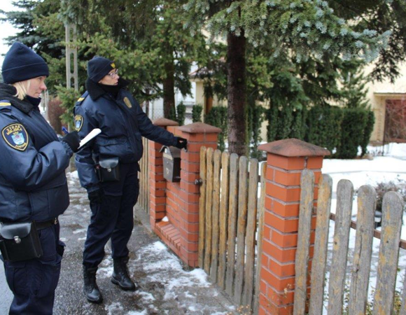 Dwie funkcjonariuszki Straży Miejskiej w granatowych mundurach i czarnych czapkach, stojące na zaśnieżonym chodniku przy drewniano-ceglanym ogrodzeniu. Funkcjonariuszka po lewej trzyma notatnik i długopis. Po prawej funkcjonariuszka wskazuje na teren posesji. W tle iglaste drzewa i budynek mieszkalny.