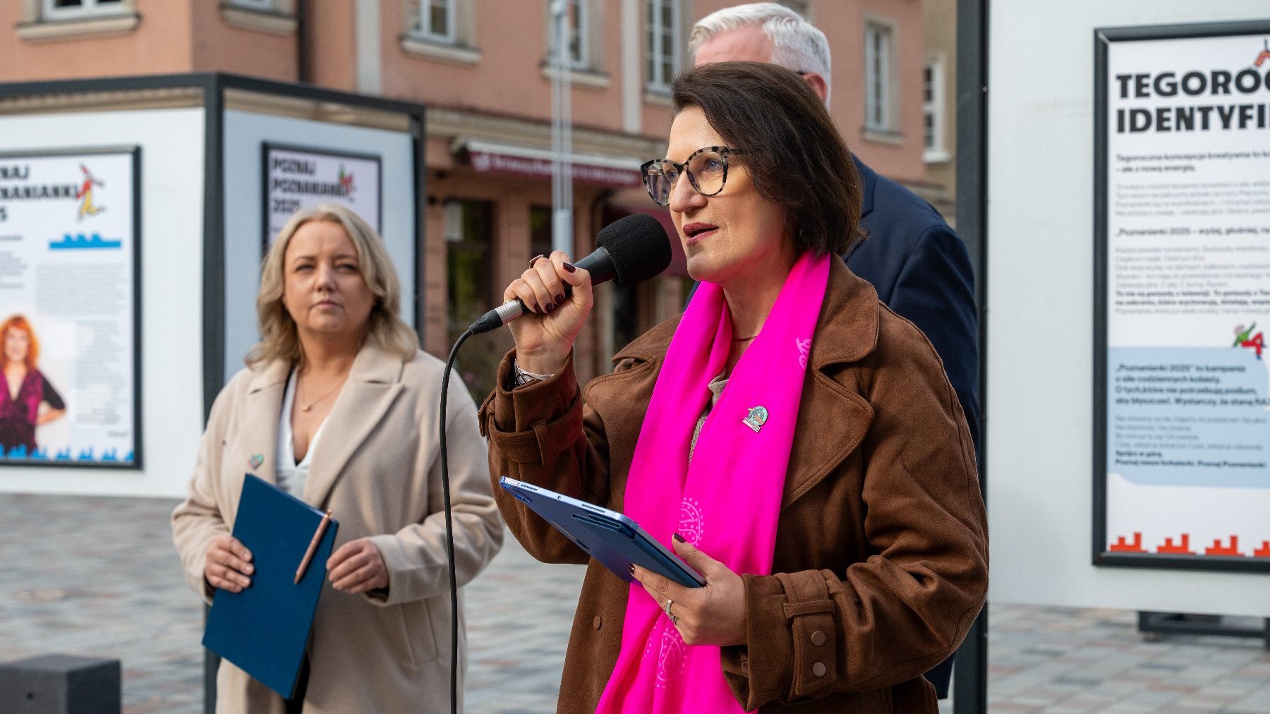 Dwie kobiety na tle wystawy na placu jedna z kobiet na pierwszym planie trzyma w ręku mikrofon za nią mężczyzna słabo widoczny na zdjęciu