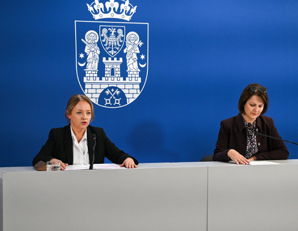 Dwie kobiety podczas konferencji prasowej za stołem prezydialnym na tle niebieskiej ścianki z herbem Poznania.
