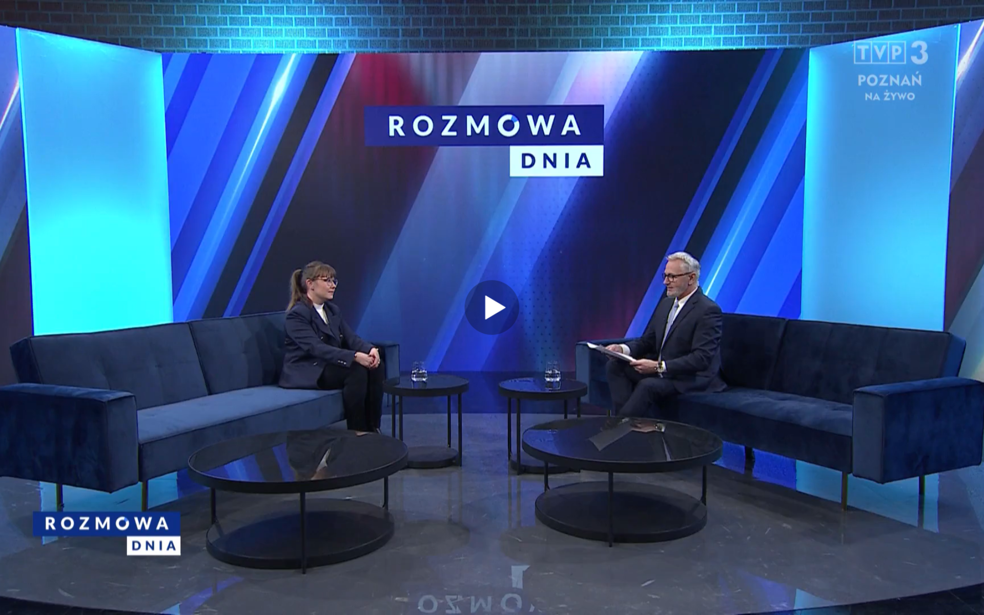 Dwie osoby siedzą na granatowych kanapach w studiu telewizyjnym programu "Rozmowa Dnia" na antenie TVP3 Poznań. Po lewej stronie kobieta w okularach, ubrana w ciemny żakiet i spodnie, z dłoniami złożonymi na kolanach. Po prawej stronie mężczyzna w garniturze, trzymający kartki. W tle nowoczesna scenografia w odcieniach niebieskiego z podświetlanymi panelami oraz logotyp programu. Na ekranie widoczne jest również oznaczenie "NA ŻYWO", wskazujące transmisję na żywo.