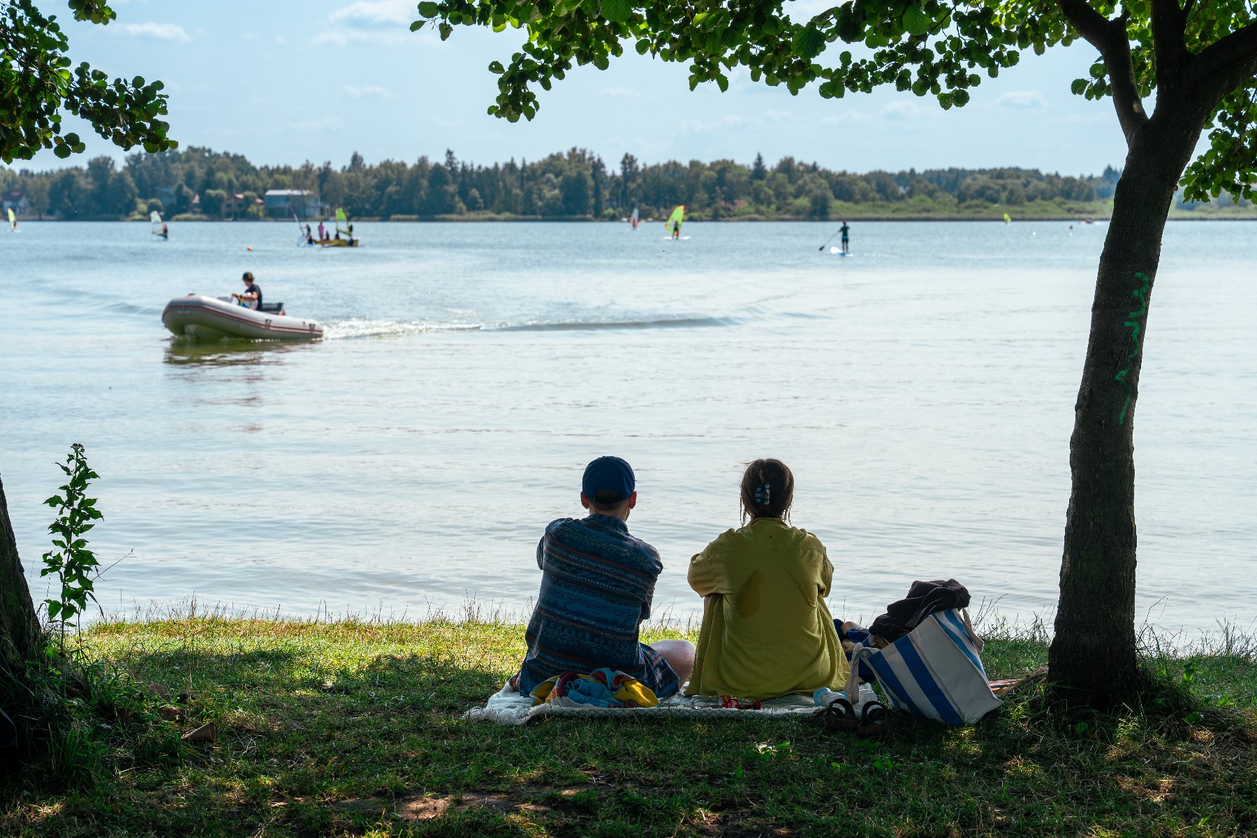 Dwie osoby siedzące na kocu na trawiastym brzegu jeziora pod cieniem drzew, obserwujące łódź motorową i windsurferów na wodzie.