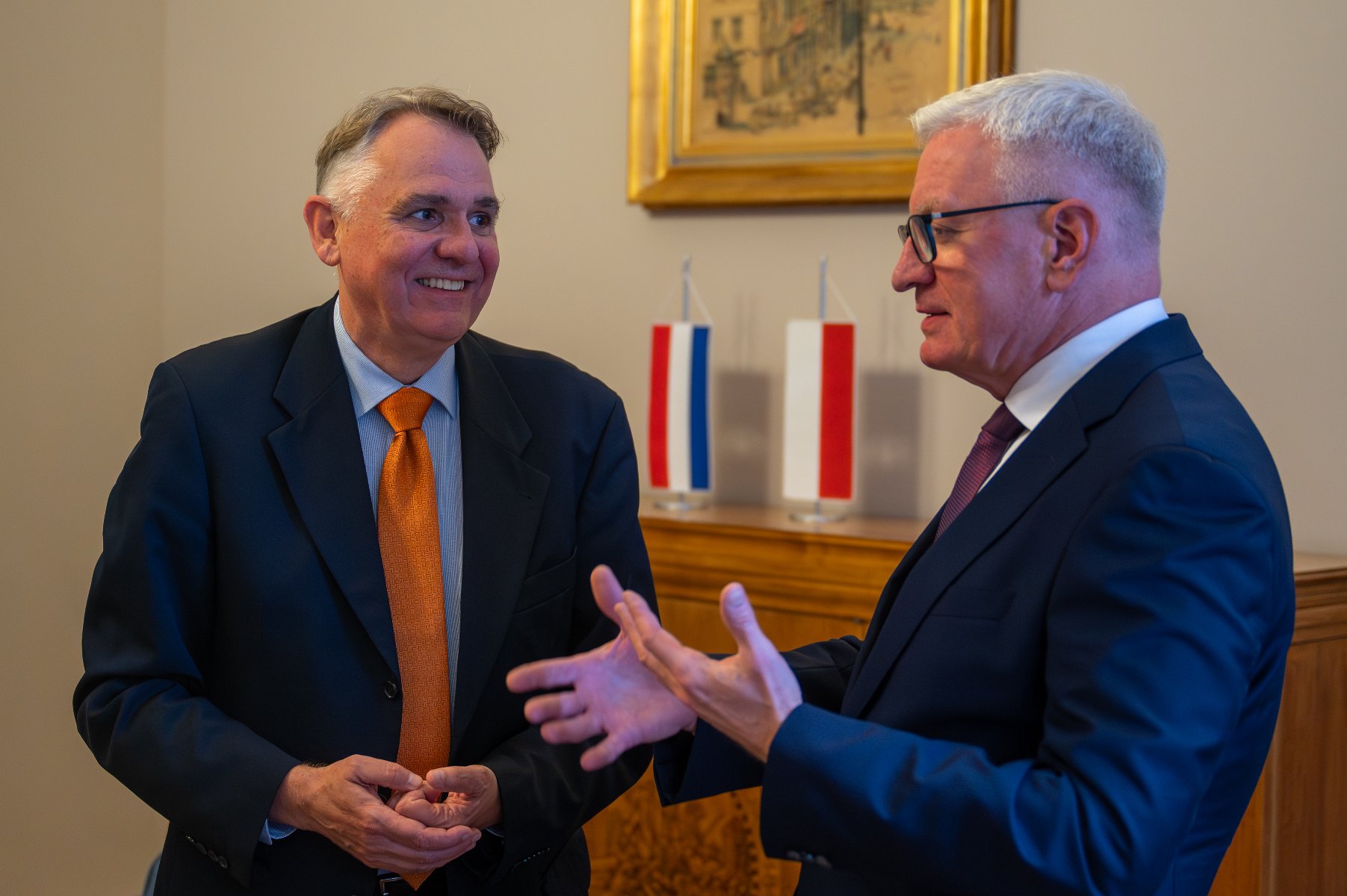 Dwóch elegancko ubranych mężczyzn, w garniturach, rozmawia w pomieszczeniu. Mężczyzna po lewej, w pomarańczowym krawacie, uśmiecha się. Mężczyzna po prawej, w okularach, gestykuluje dłońmi. W tle widać dwie małe flagi: jedną biało-czerwono-niebieską i drugą biało-czerwoną.