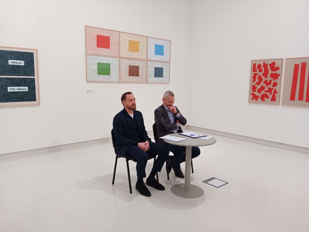 Dwóch mężczyzn w garniturach siedzi przy okrągłym stoliku, za nimi obrazy z wystawy muzealnej.