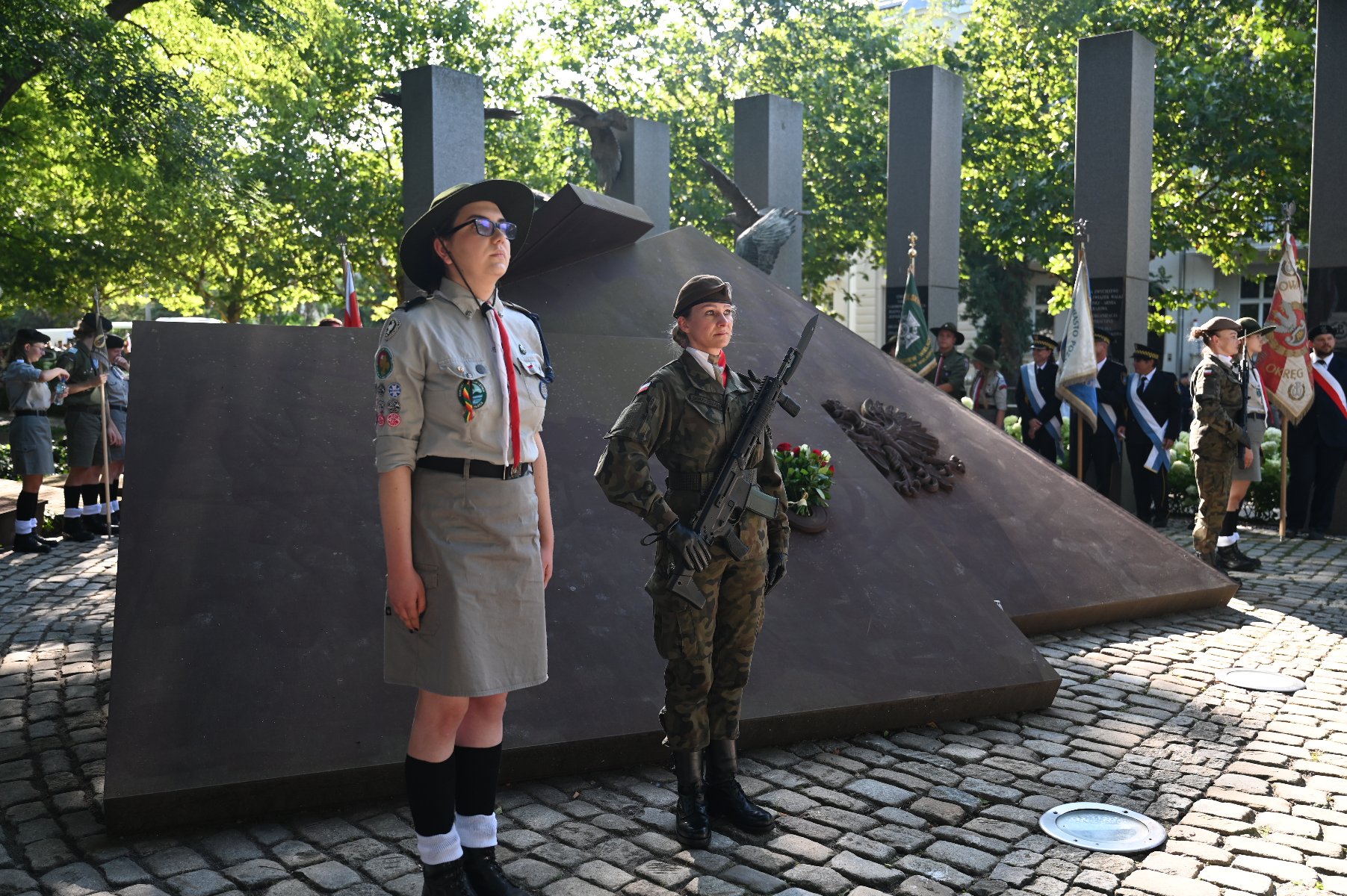 Dwóch młodych ludzi w mundurach stoi na baczność przed pomnikiem. Po lewej harcerka w jasnym mundurze i kapeluszu, a po prawej żołnierka w mundurze polowym z karabinem i kwiatami. Za nimi widoczny jest masywny, ciemny pomnik, a w tle zieleń drzew i inne postacie w mundurach, oraz flagi. Scena wygląda jak uroczystość patriotyczna.
