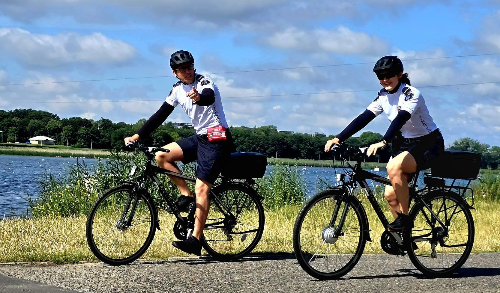Dwóch rowerzystów, prawdopodobnie policjantów lub strażników miejskich, w czarnych kaskach i białych koszulkach z długimi rękawami, jedzie na rowerach po ścieżce rowerowej. Obok nich znajduje się jezioro lub rzeka, a w tle widać zieleń i błękitne niebo z chmurami. Mężczyzna po lewej stronie wskazuje palcem w kierunku widza.