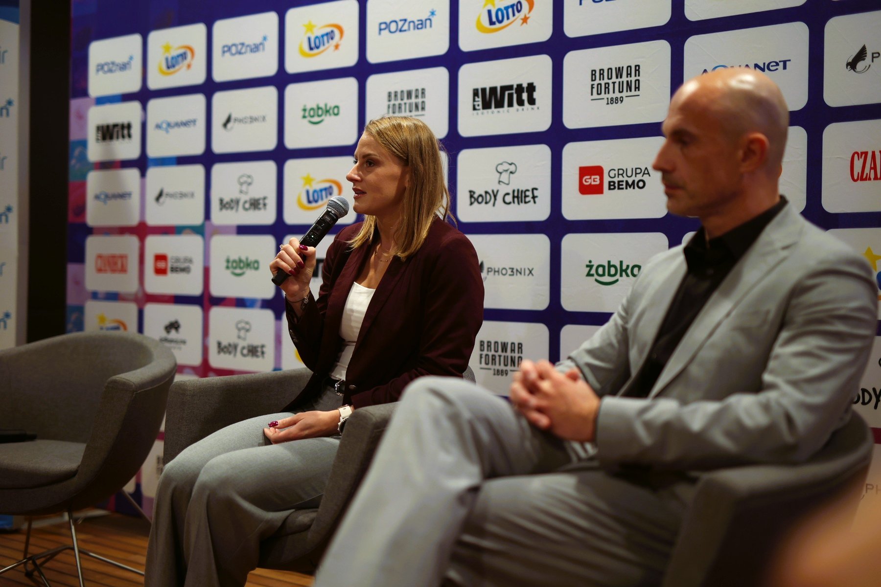 Dwóch uczestników konferencji prasowej LOTTO 24. Poznań Maratonu - kobieta przemawia i trzyma mikrofon, a mężczyzna siedzi w garniturze