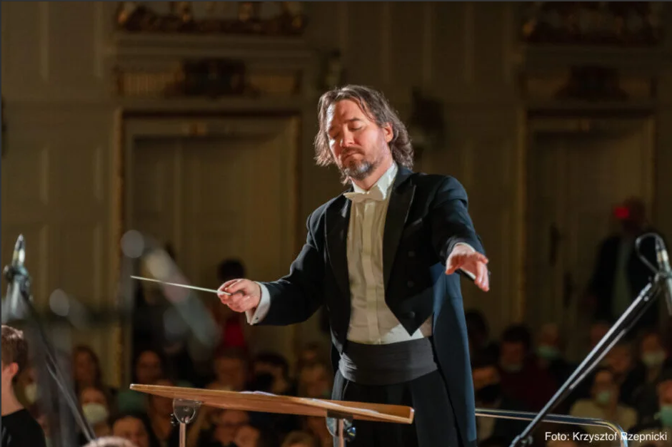 Dyrygent w eleganckim fraku prowadzący orkiestrę podczas koncertu w zabytkowej sali. W tle widoczna publiczność i ciepłe, sceniczne oświetlenie.