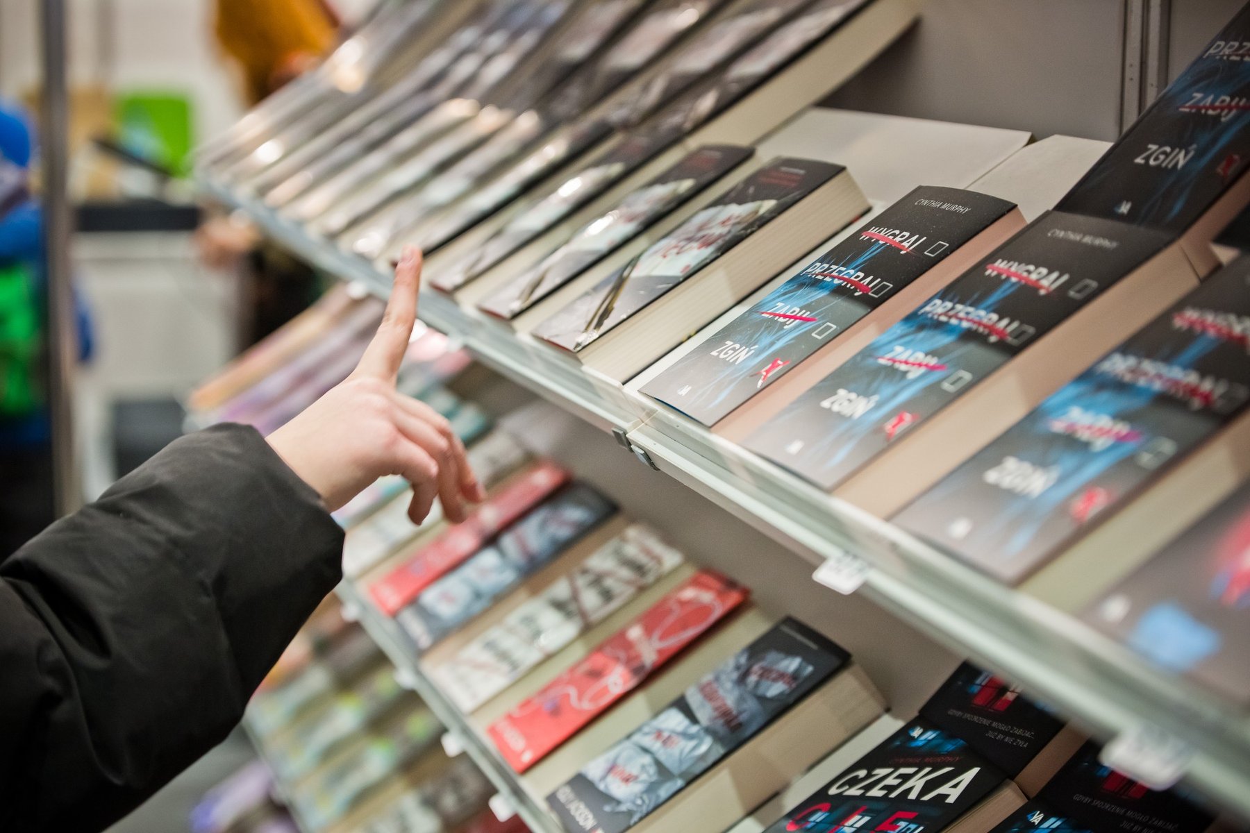 Dzecięca ręka wskazuje palcem na półkę z poukładanymi książkami. Książki leżą okładką skierowaną do góry.