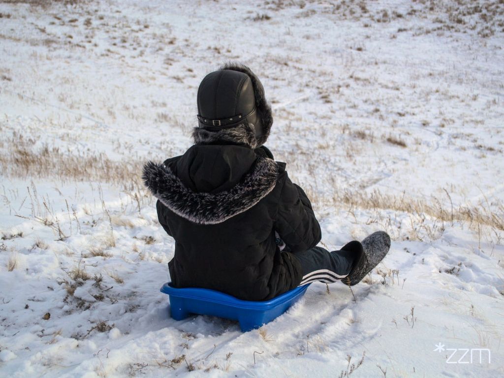 Dziecko w czarnej, pikowanej kurtce z futrzanym kapturem oraz czarnej czapce uszance siedzi tyłem, zjeżdżając na małych, niebieskich sankach po zaśnieżonym, lekko pochyłym stoku