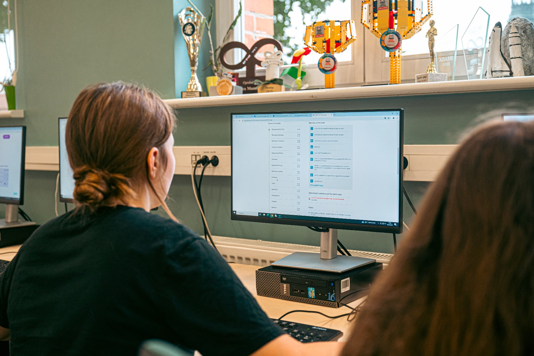 Dziewczyna siedzi tyłem do kamery przed monitorem komputerowym, pracując. Na ekranie widać otwartą aplikację. Na biurku obok komputera leży klawiatura. W tle, na parapecie, stoją liczne puchary i trofea, w tym model promu kosmicznego i statuetka robota z klocków.