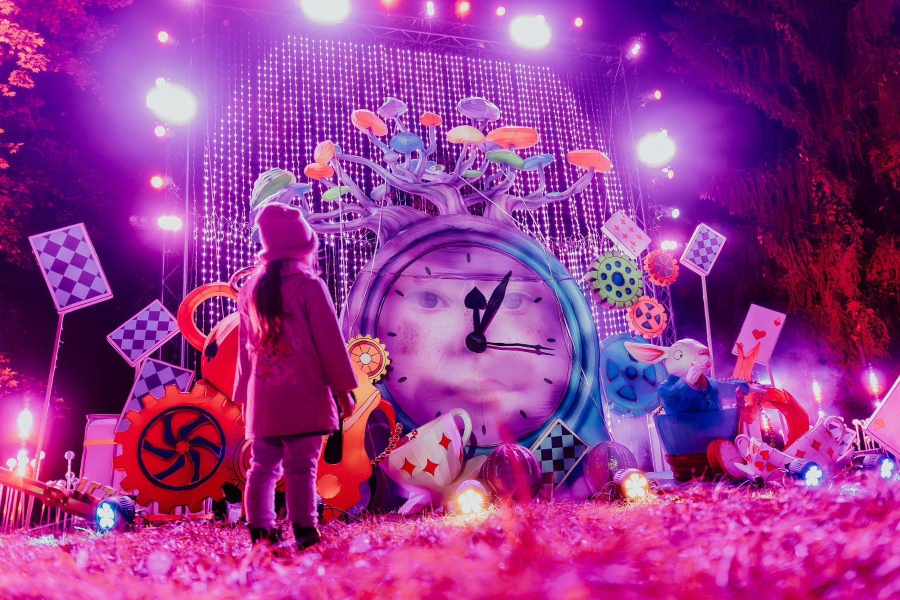 Dziewczynka ubrana w zimowy płaszcz i czapkę stoi przed świetlną instalacją, która przedstawia przedmioty z bajki Alicja w Krainie Czarów. Wokół dużego zeraga rosną kolorowe grzyby, wiszą karty i siedzi Biały Królik.