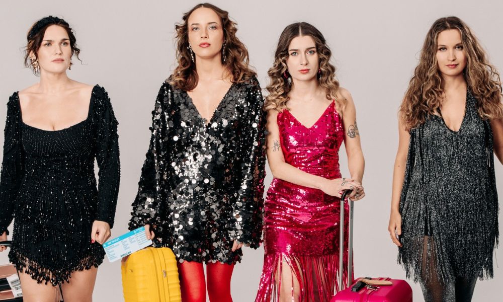 Dziewczyny leżą stykając się głowami, korzystają z telefonów, jedna robi sobie selfie, druga rozmawia, trzecia coś sprawdza.