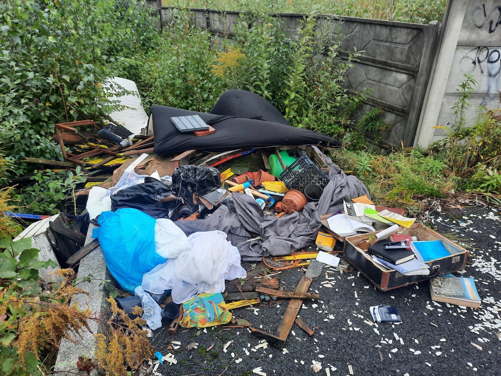Dzikie wysypisko śmieci na poboczu. Na środku kadru leży duży stos odpadów, w tym czarny materac, deski, ubrania, torby, plastikowe pojemniki i książki. W tle betonowy mur, porośnięty wysokimi chwastami.