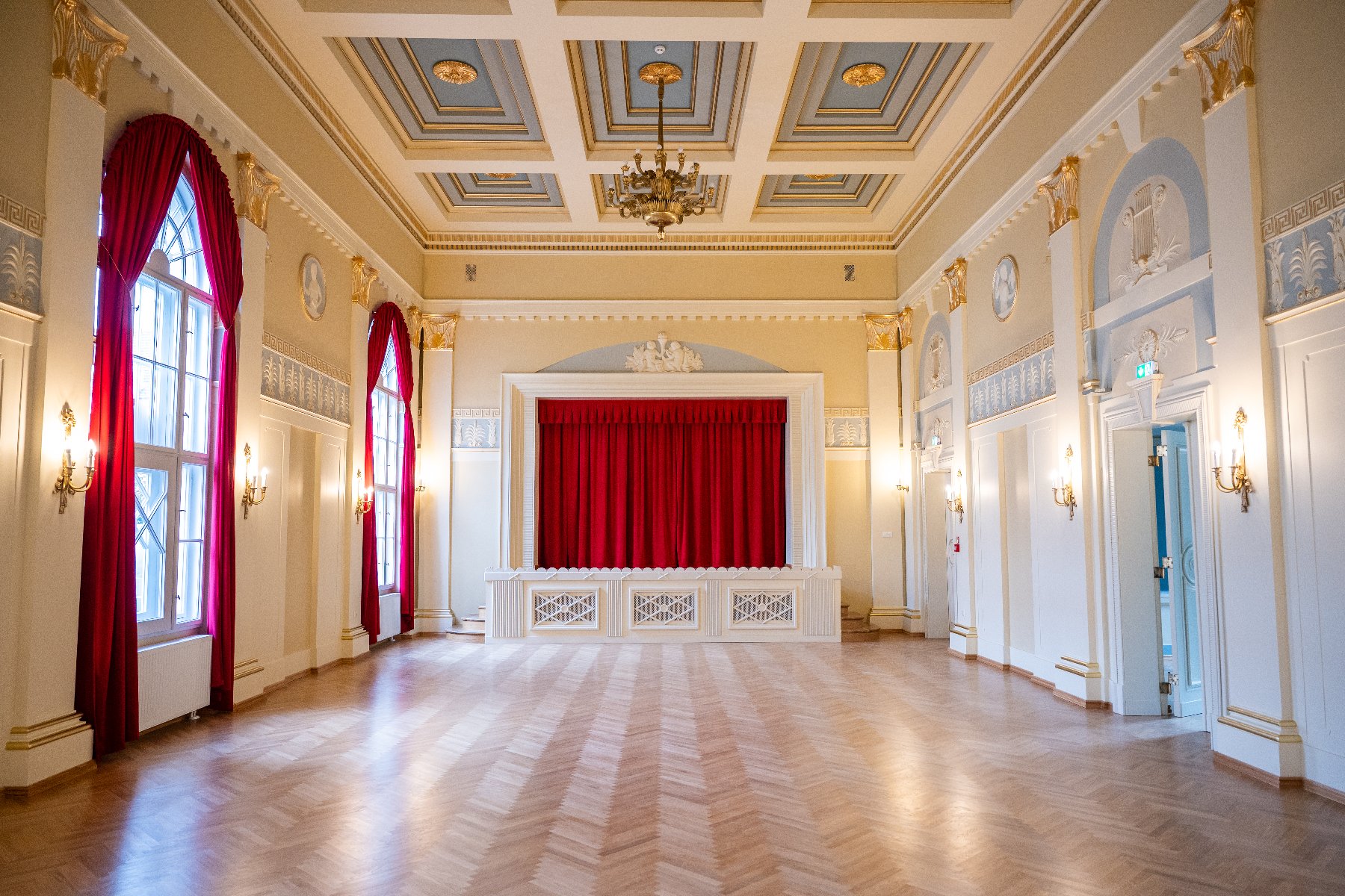 Eleganckie, puste wnętrze reprezentacyjnej sali balowej z parkietem w jodełkę, zdobionym kasetonowym sufitem i sceną z czerwoną kurtyną.