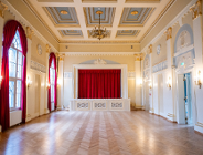 Eleganckie, puste wnętrze reprezentacyjnej sali balowej z parkietem w jodełkę, zdobionym kasetonowym sufitem i sceną z czerwoną kurtyną.