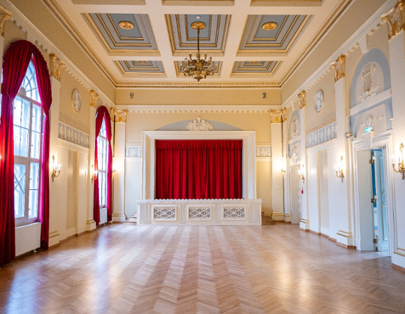 Eleganckie, puste wnętrze reprezentacyjnej sali balowej z parkietem w jodełkę, zdobionym kasetonowym sufitem i sceną z czerwoną kurtyną.