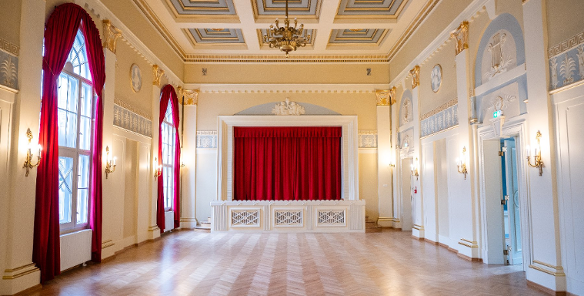 Eleganckie, puste wnętrze reprezentacyjnej sali balowej z parkietem w jodełkę, zdobionym kasetonowym sufitem i sceną z czerwoną kurtyną.