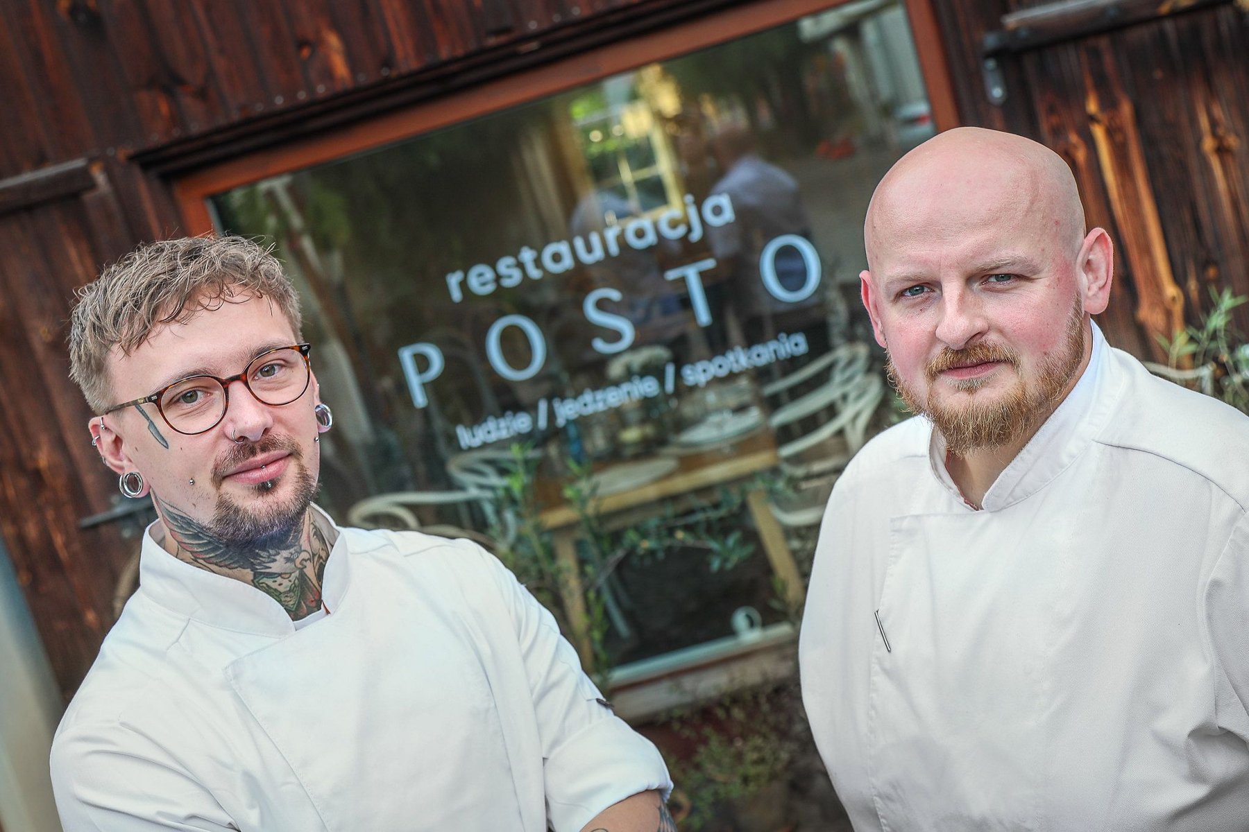 Eleganckie wnętrze restauracji sushi. Na pierwszym planie owalny stół z dużą donicą, w której rośnie palma. Na stołach i przy barze widoczne naczynia i pałeczki. W tle rzędy stolików i krzeseł, a także regały z dekoracjami. Duże okna wpuszczają światło dzienne.