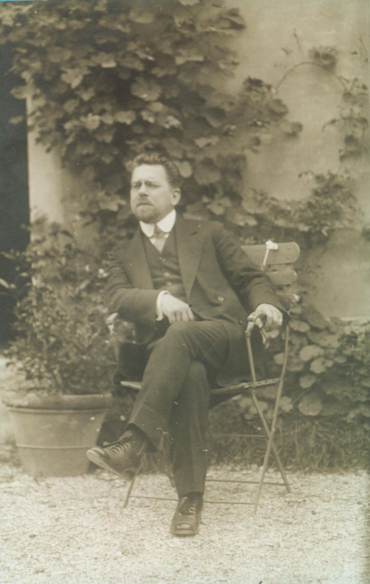 Elegancko ubrany mężczyzna siedzi na krześle pod domem, wśród zieleni.