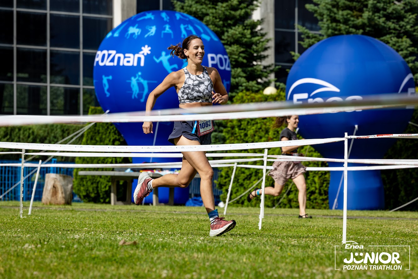 Enea Junior Poznań Triathlon 2024, fot. Jarosław Koperski - uśmiechnięta kobieta biegnie po trawie na tle niebieskich balonów Poznania i Enei. W oddali widać także inną biegnącą kobietę