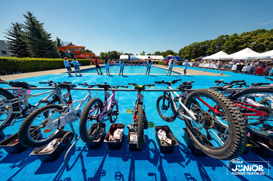 Enea Junior Poznań Triathlon 2026, fot. Enea Junior Poznań Triathlon - na pierwszym planie rowery dziecięce, w tle osoby pomagające wyjść dzieciom z wody, basen, a po prawej stronie rodziny zawodników