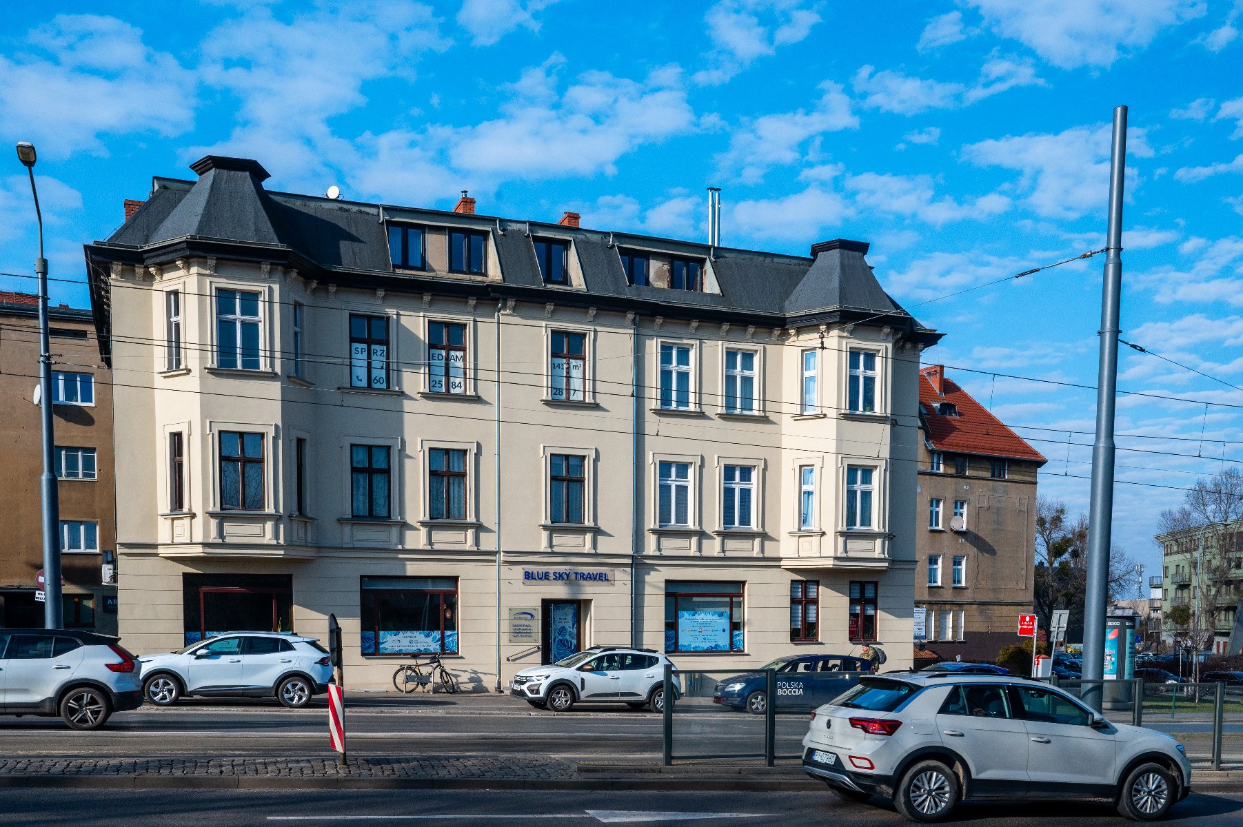 Fasada beżowego budynku z architektonicznymi detalami, takimi jak dekoracyjne listwy i gzymsy. Budynek ma duże okna z białymi ramami, a niebo jest jasne z kilkoma chmurami. Perspektywa zdjęcia jest z niskiego kąta, patrzącego w górę na budynek, co podkreśla wysokość i cechy architektoniczne.