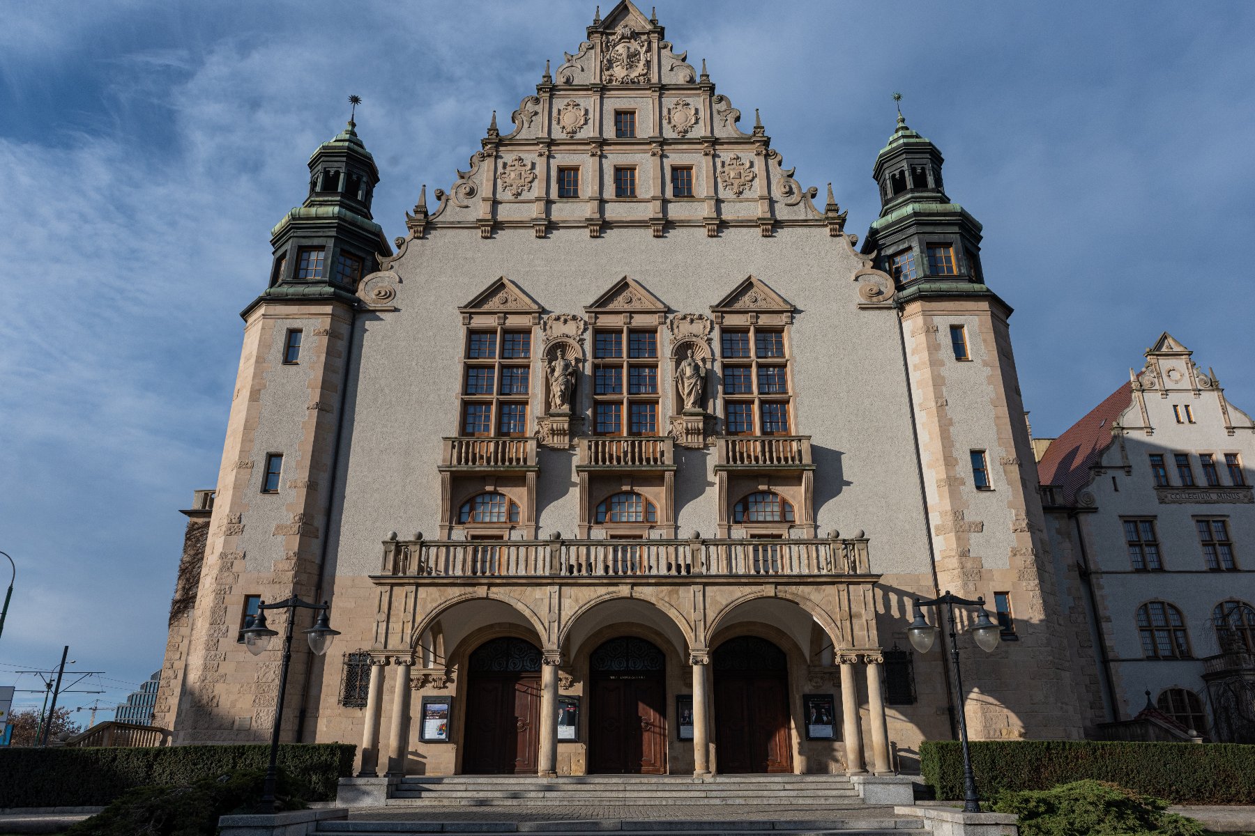 Fasada budynku Uniwersytetu im. Adama Mickiewicza w Poznaniu, z charakterystyczną ozdobną attyką, dwoma wieżami po bokach i rzeźbami nad oknami. Budynek posiada liczne okna i drzwi, a przed nim znajdują się schody prowadzące do wejścia.