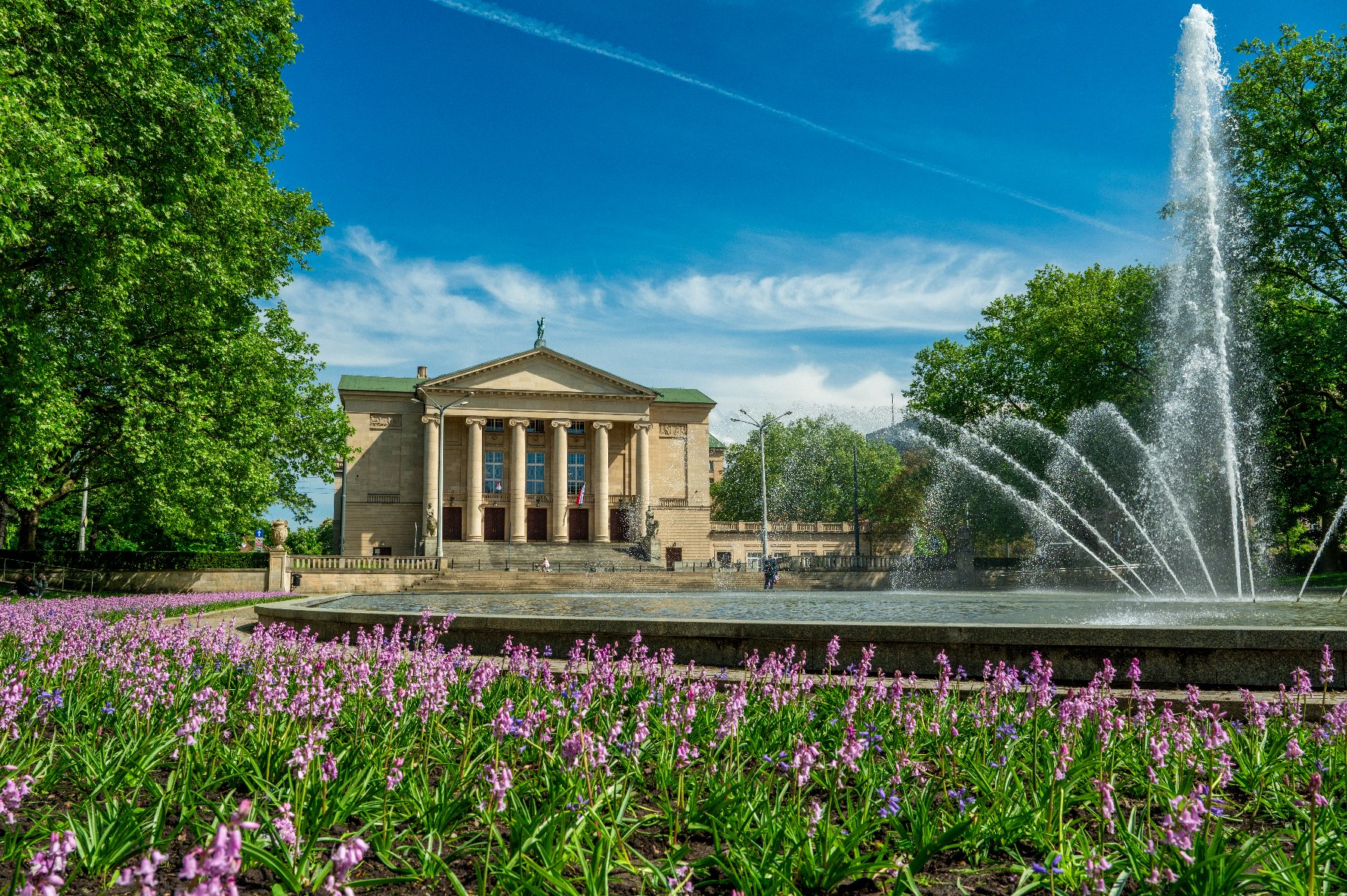 Fasada Teatru Wielkiego im. Stanisława Moniuszki w Poznaniu, otoczonego zielonym parkiem z kwitnącymi, liliowymi kwiatami na pierwszym planie. Przed teatrem znajduje się duża fontanna tryskająca wodą. Błękitne niebo z lekkimi smugami chmur dopełnia obraz.