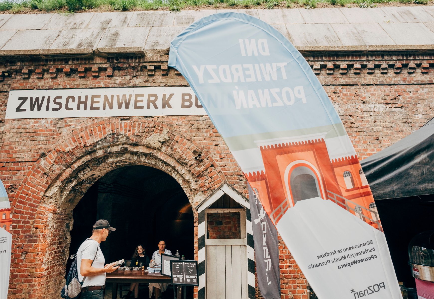 Flaga promocyjna wydarzenia Dni Twierdzy Poznań przed wejściem do zabytkowego Fortu Va i punktem informacyjnym.