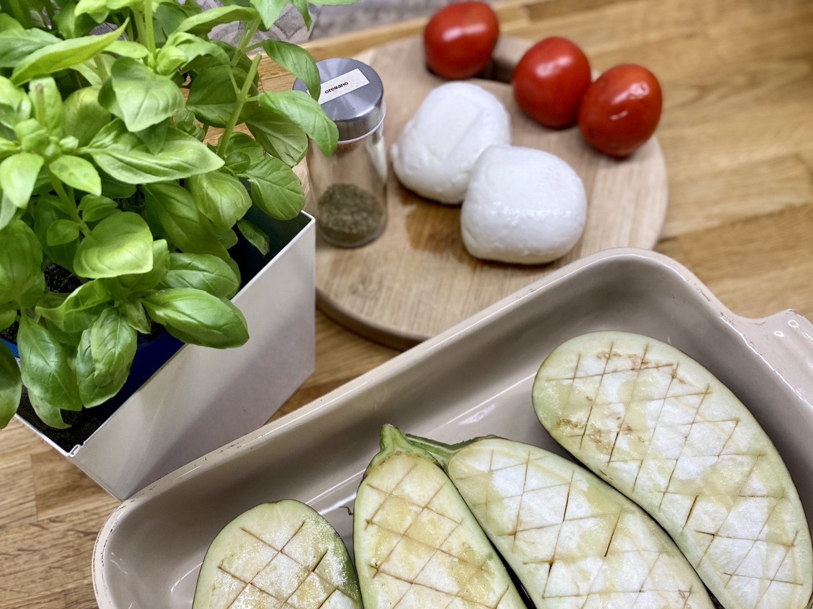 Foremka z bakłażanami naciętymi w kratkę, obok kulki mozzarelli, pomidory i bazylia.