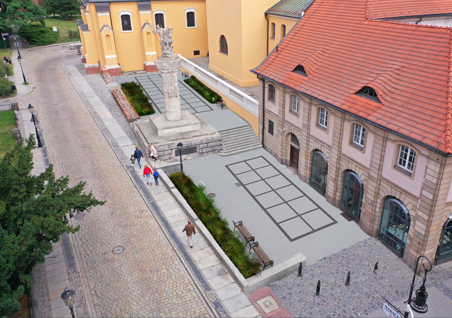 Fotografia przedstwiająca widok na pomnik opisany w tekście. Widok z lotu ptaka. Pomnik na tle budynków kościoła i kamienicy z czerwonym dachem.