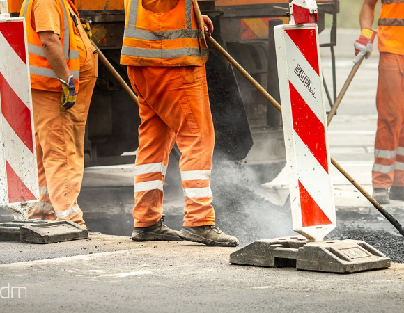 Fragment budowy drogi. Trzech pracowników ubranych w pomarańczowe spodnie i kamizelki odblaskowe stoi na asfalcie, trzymając szpadle lub grabie do wyrównywania nawierzchni. Na pierwszym planie dwa biało-czerwone słupki drogowe. Z lewej strony na dole widoczne małe, białe logo w cudzysłowie: "*zdm".