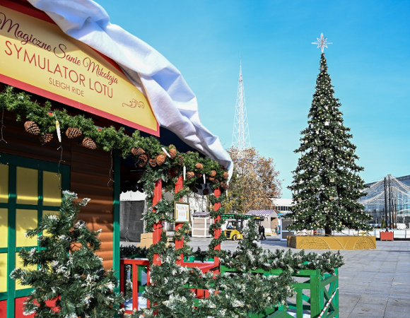 Fragment drewnianego, świątecznego stoiska z białym dachem i dekoracjami z zielonych gałązek i szyszek. Na daszku czerwony napis: "Magiczne Sanie Mikołaja SYMULATOR LOTU SLEIGH RIDE". W tle duża, udekorowana choinka z gwiazdą na szczycie i biała, spiczasta wieża radiowa na tle błękitnego nieba.