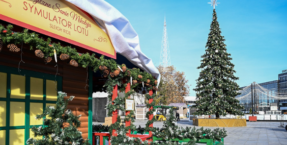 Fragment drewnianego, świątecznego stoiska z białym dachem i dekoracjami z zielonych gałązek i szyszek. Na daszku czerwony napis: "Magiczne Sanie Mikołaja SYMULATOR LOTU SLEIGH RIDE". W tle duża, udekorowana choinka z gwiazdą na szczycie i biała, spiczasta wieża radiowa na tle błękitnego nieba.