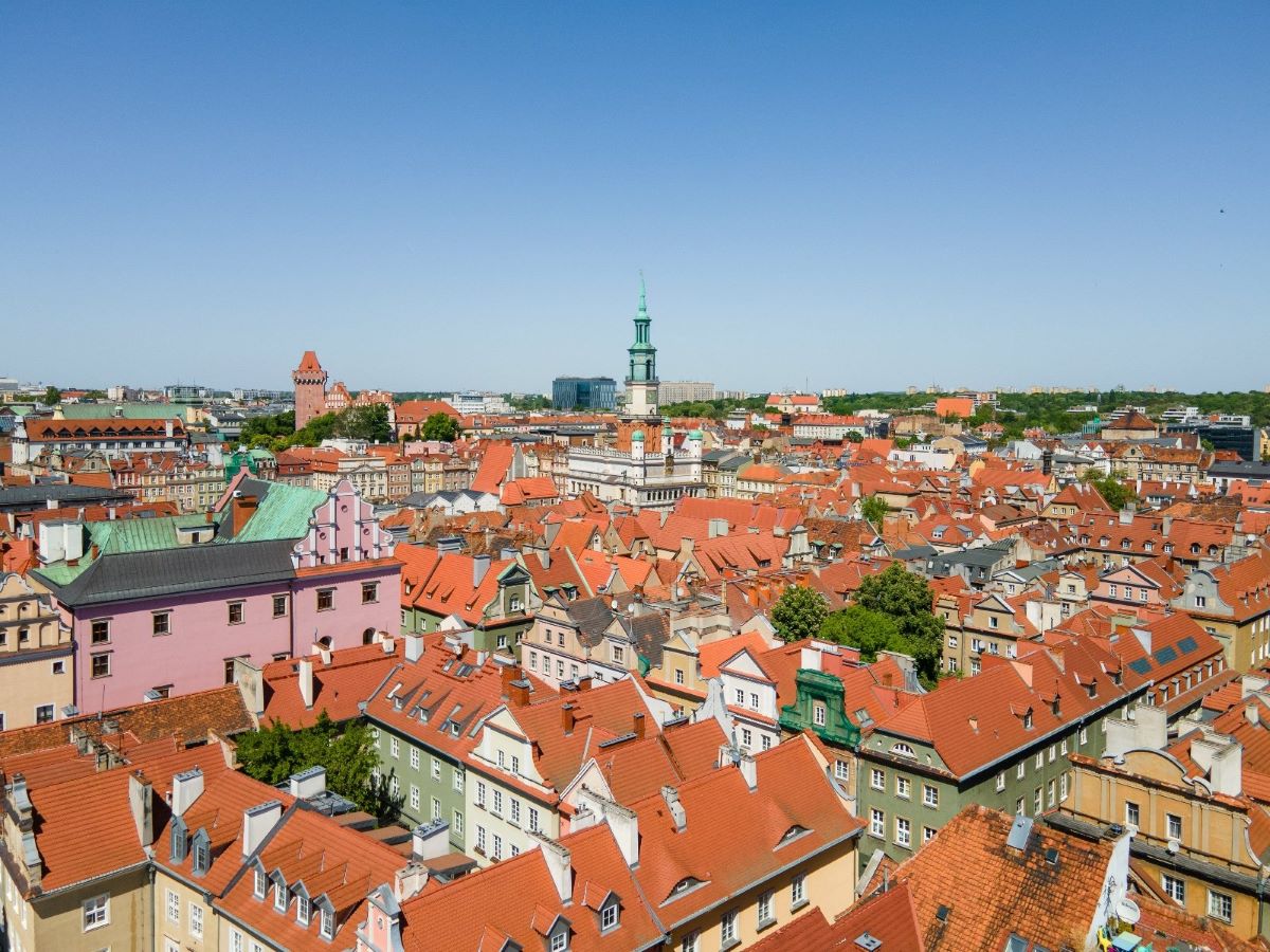 fragment panoramy Starego Miasta w Poznaniu, widoczne dachy kamienic z czerwoną dachówką, w tle widoczna wieża ratusza i wieża zamku Przemysła, na miastem błękitne niebo, słoneczna pogoda