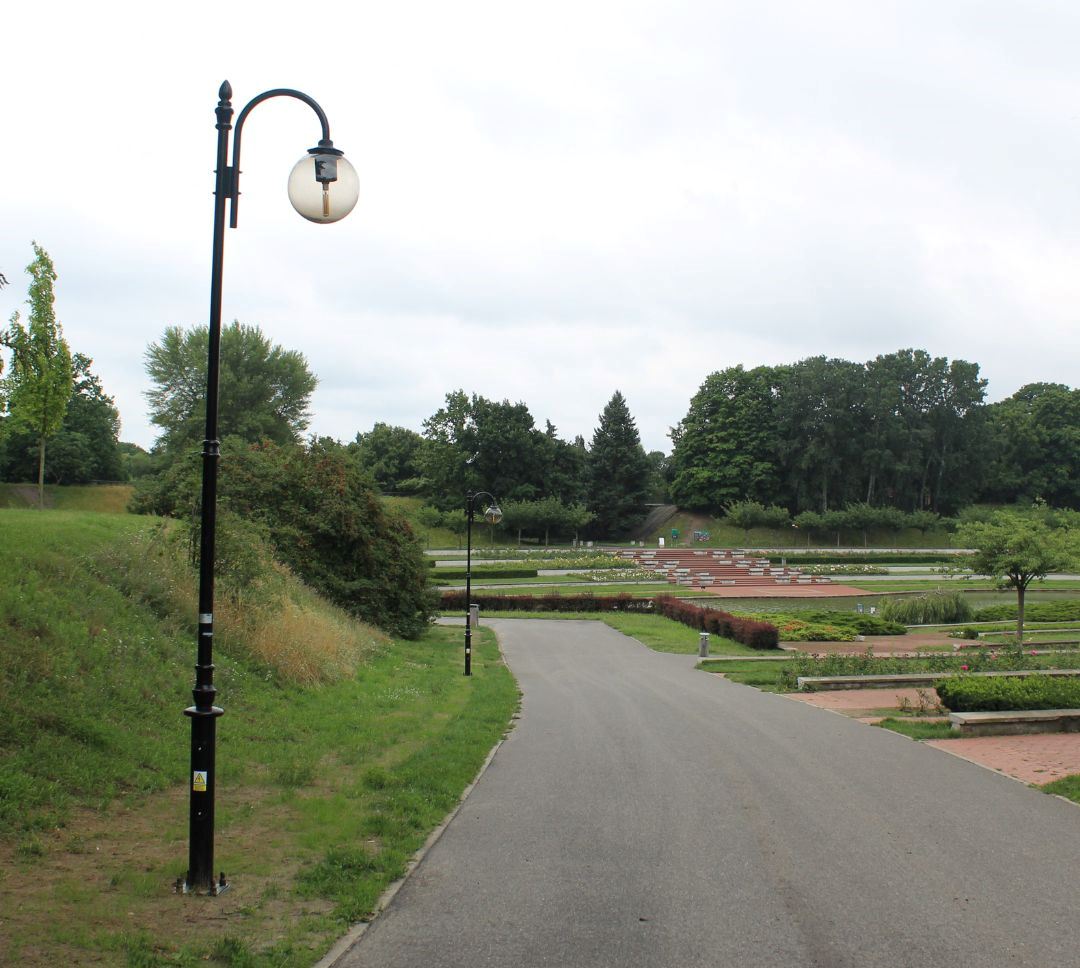 fragmnet parku z asfatową ścieżką wiodącą do oczka wodnego otoczonego ławkami, kwiatami, niską ziesilenią, przy alejce lampy uliczne, w dalszej części wysokie drzewa