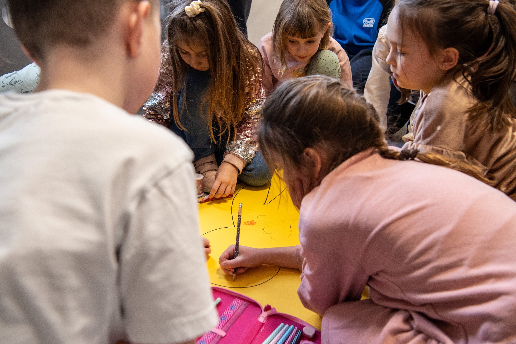 Grupa dzieci siedzi na podłodze i rysuje na dużym żółtym arkuszu papieru. Na pierwszym planie piórnik z kredkami i ołówkami. Dzieci pochylone nad pracą, skupione na wspólnym rysunku.