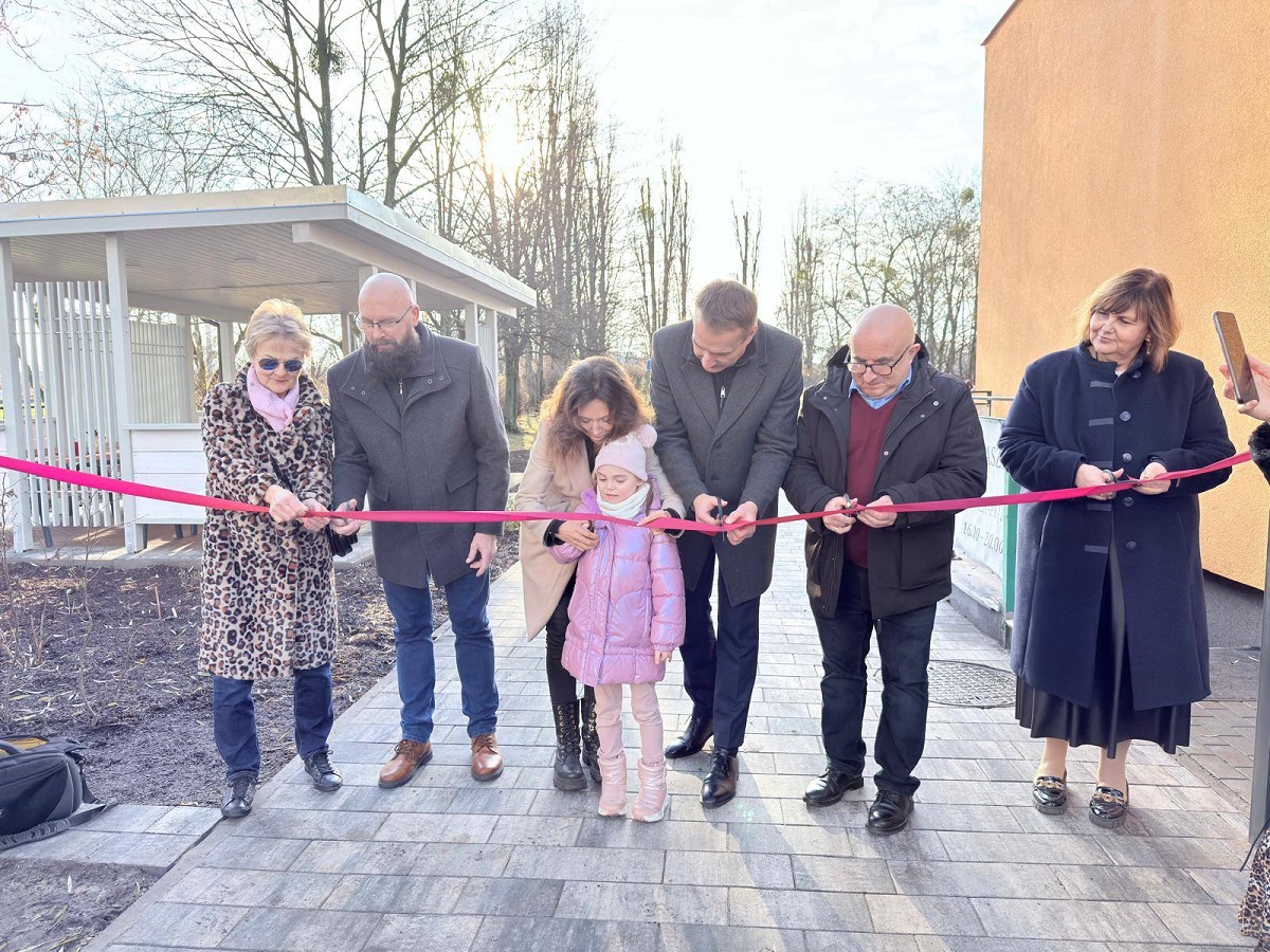 grupa kilkorga osób kobiet, mężczyzn, dziecko przecinaja wstęgę na terenie przed budynkiem przedszkola