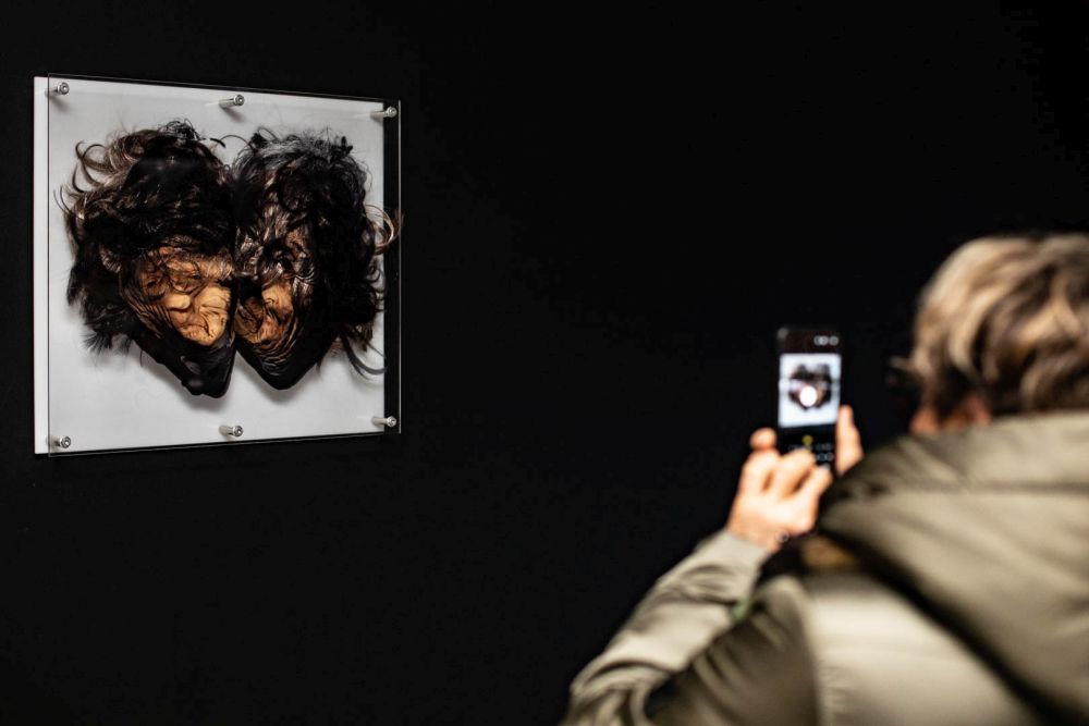 Grupa ludzi przygląda się wielu małym fotografiom wiszącym na białej ścianie.