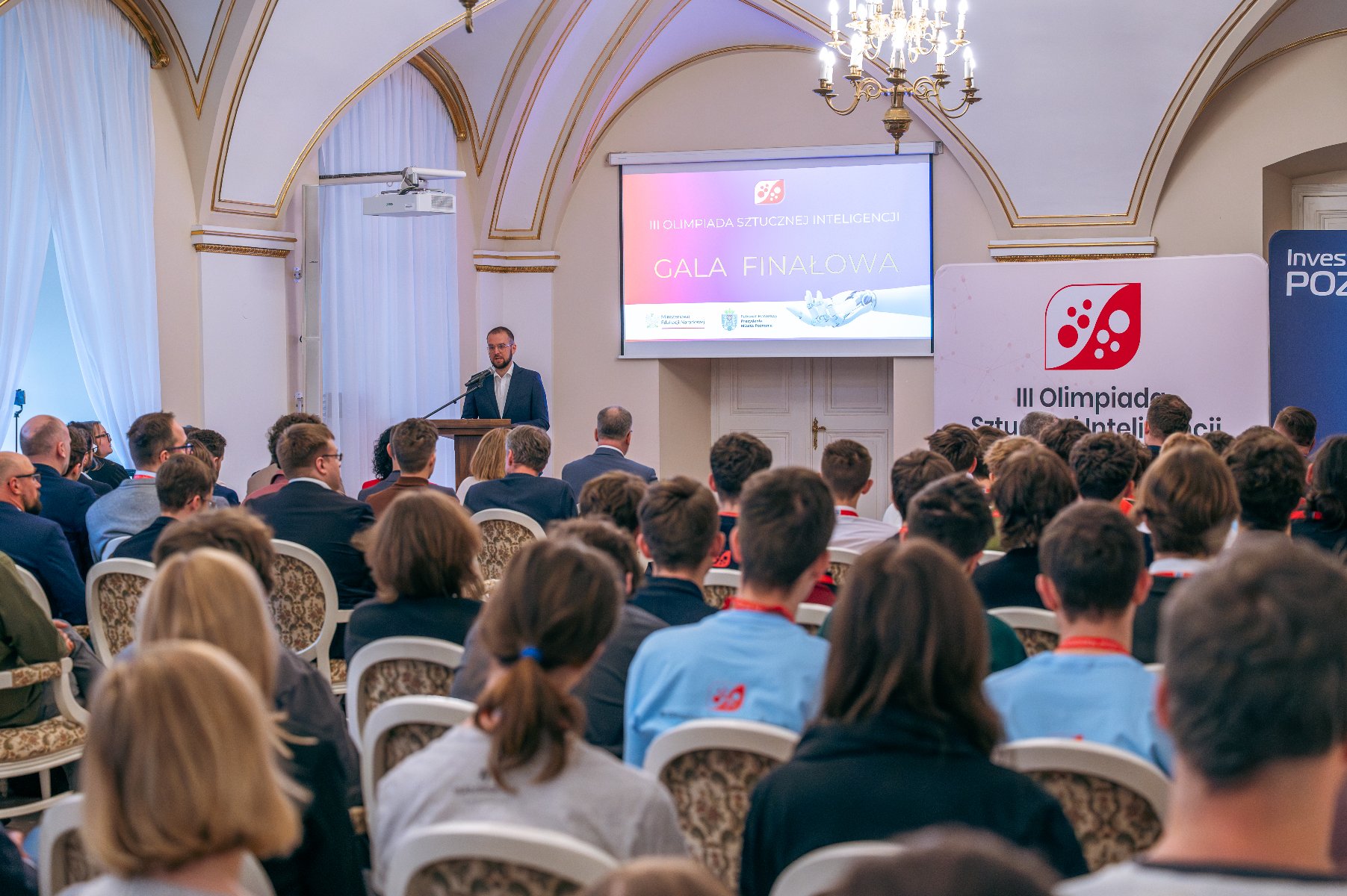 Grupa ludzi siedząca w eleganckiej sali, przy mikrofonie stoi mężczyzna. Na rzutniku pojawia się napis III Olimpiada Sztucznej Inteligencji, obok stoją dwa roll-upy