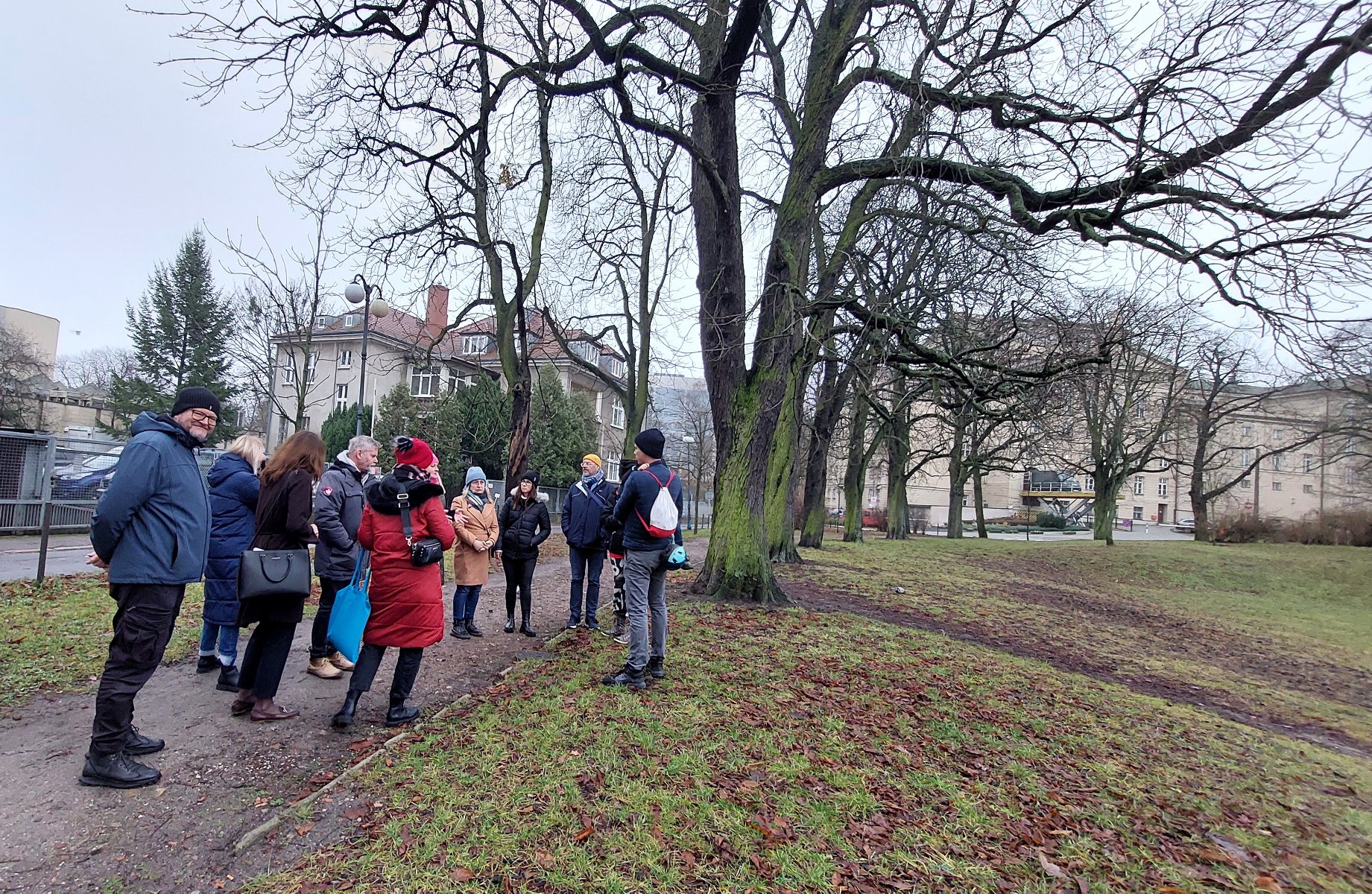 Grupa ludzi spaceruje po Parku Wieniawskiego.Widać, że rozmawiają. Na drugim planie widać wysokie drzewa, a w oddali budynki