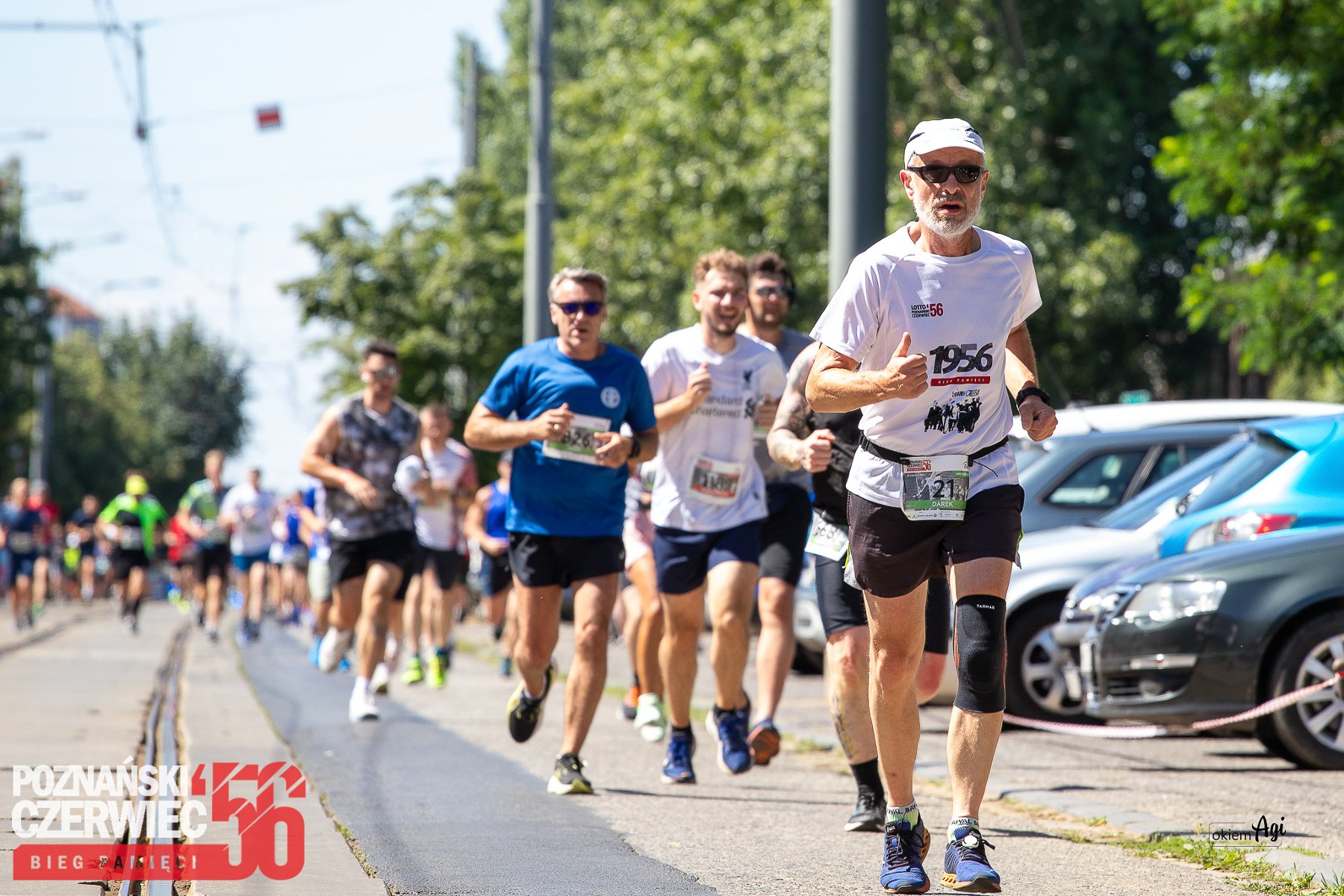 Grupa mężczyzn biegnie w stronę obiektywu. Są w różnym biegu i ubrani w stroje sportowe. W tle samochody, drzewa i reszta biegaczy.