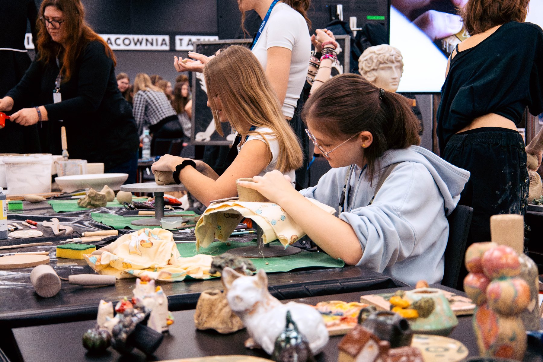 Grupa młodych osób siedzi przy długim stole i lepi z gliny w pracowni ceramicznej; na blacie widać narzędzia, gotowe figurki oraz miski z materiałami.