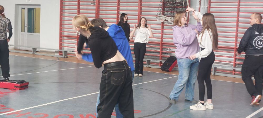 Grupa młodzieży uczestniczy w zajęciach samoobrony na sali gimnastycznej. Uczniowie, w parach, ćwiczą techniki obrony pod okiem instruktora. Na pierwszym planie dwie pary intensywnie trenują, a w tle widać czerwone drabinki gimnastyczne.